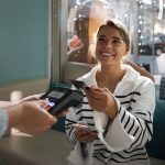 A smiling woman in a cozy café uses her credit card to make a payment while holding her phone, representing customer engagement and the use of loyalty programs to enhance shopping experiences.