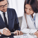 Two business professionals discussing finances on a digital tablet during a meeting, with printed charts and reports on the table.