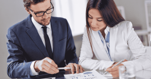 Two business professionals discussing finances on a digital tablet during a meeting, with printed charts and reports on the table.