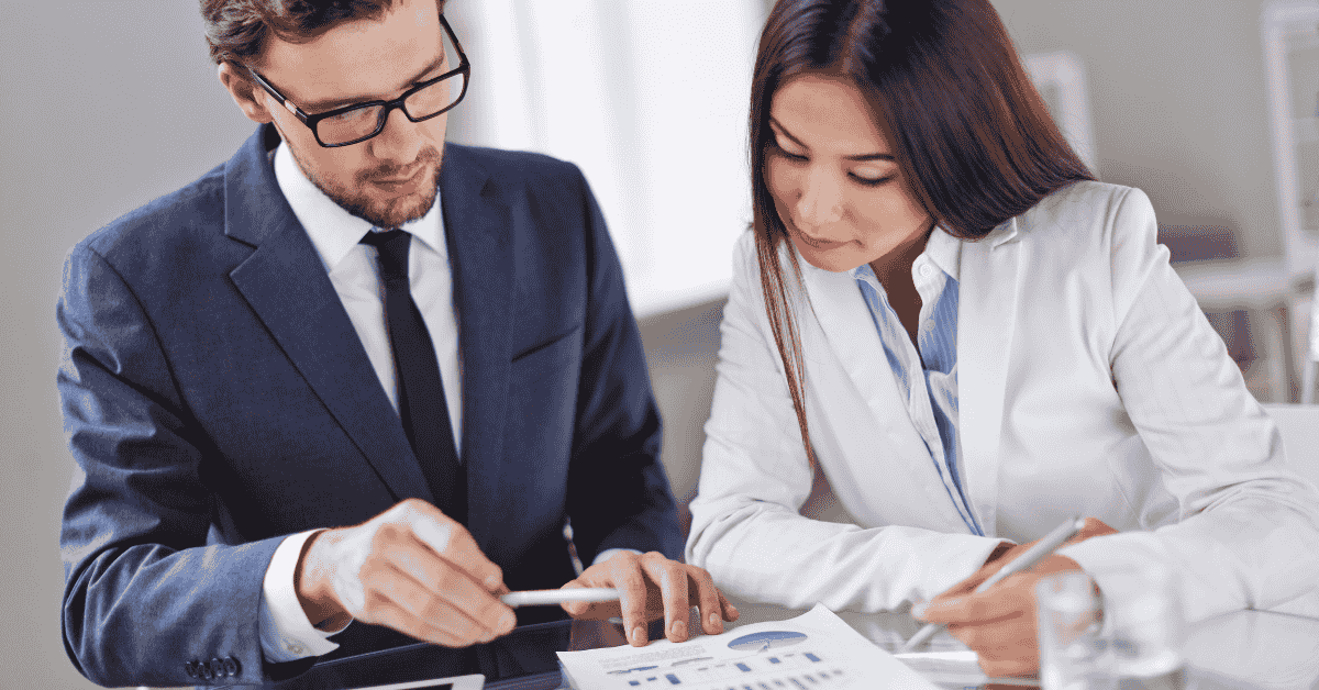 Two business professionals discussing finances on a digital tablet during a meeting, with printed charts and reports on the table.