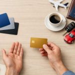 Hands holding a gold credit card over a desk with laptop, coffee, passport, toy car, and salad bowl.