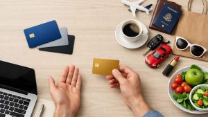 Hands holding a gold credit card over a desk with laptop, coffee, passport, toy car, and salad bowl.